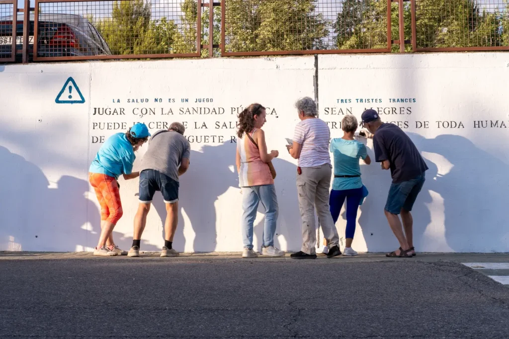 Seis adultos permanecen de pie junto a una pared blanca con escritura, algunos leen poesías y otros interactúan con el texto. Al fondo se ven árboles y una valla metálica. El soleado escenario al aire libre sugiere un acto comunitario en Rivas.