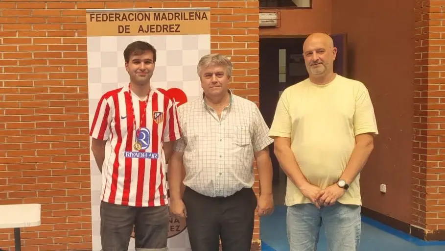 Tres hombres están de pie en el interior, delante de un cartel en el que se lee «Federación Madrileña de Ajedrez». Uno lleva una camiseta deportiva a rayas rojas y blancas, y los otros dos llevan camisetas de colores claros. Detrás de ellos se ve una pared de ladrillo.