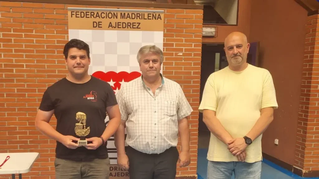 Tres hombres están de pie en el interior, frente a un cartel en el que se lee «Federación Madrileña de Ajedrez». El hombre de la izquierda sostiene un trofeo, mientras que los otros dos mantienen una expresión impasible. Al fondo se ve una pared de ladrillo.