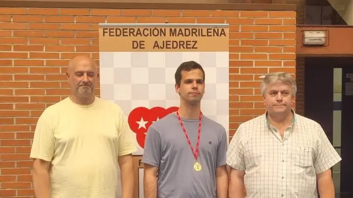 Tres hombres posan delante de un cartel de la Federación Madrileña de Ajedrez. El hombre del centro lleva una medalla de oro y una camiseta gris, mientras que los otros dos se sitúan a ambos lados de él, todos mirando a la cámara.