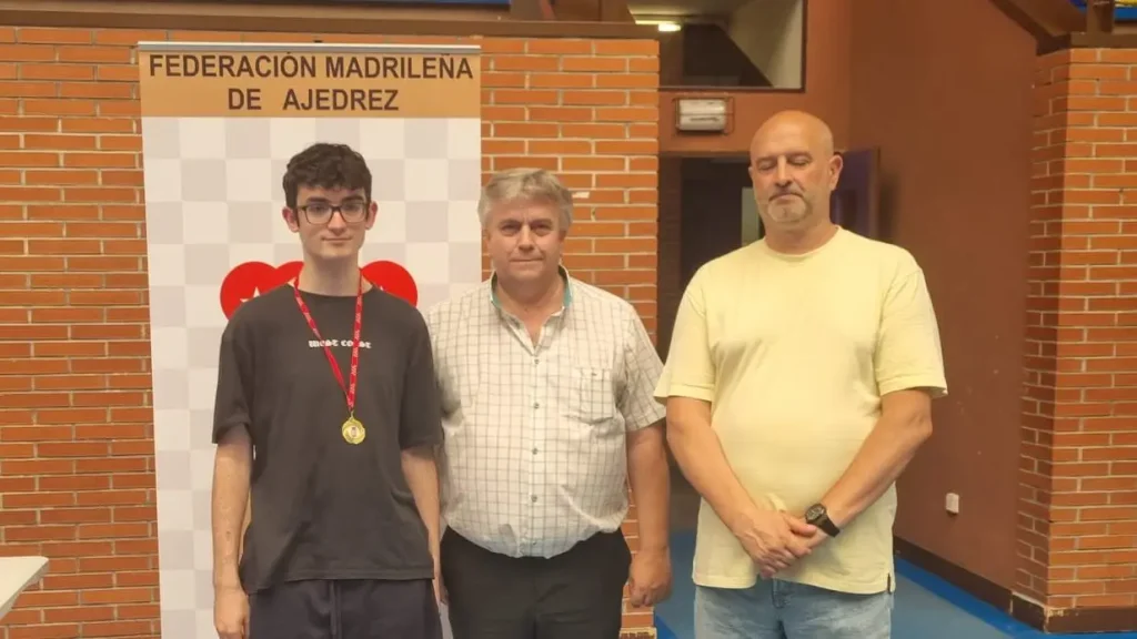 Tres hombres posan en el interior, delante de un cartel de la Federación Madrileña de Ajedrez. El hombre de la izquierda lleva una medalla y gafas; el del centro, una camisa a cuadros; y el de la derecha, una camiseta amarilla.