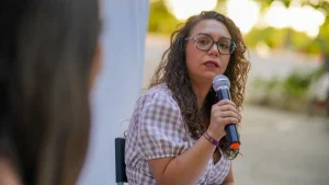 Aída Castillejo, una mujer de pelo rizado y con gafas, sostiene un micrófono y habla mientras está sentada al aire libre. Lleva una blusa estampada y mira hacia otra persona cuya espalda se ve en primer plano.