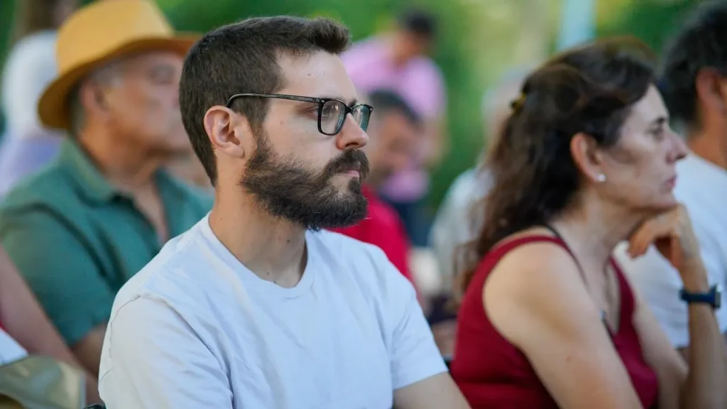 Un hombre con gafas y barba, vestido con una camiseta blanca, está sentado entre un grupo de personas al aire libre en Rivas. La mayoría de los presentes miran hacia delante y parecen estar escuchando con atención, quizá hablando de temas como la reelección o Aída Castillejo.