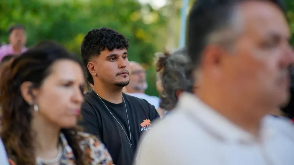 Un joven con una camiseta negra se encuentra de pie entre un grupo de personas al aire libre en Rivas, mirando al frente con expresión impasible. El fondo es verde y está desenfocado, mientras que en primer plano se ve parcialmente a otras personas, que quizá estén hablando de la reelección.