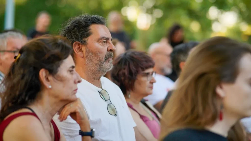 Un grupo de adultos está sentado muy juntito al aire libre, escuchando con atención algo —quizás una charla de Aída Castillejo sobre la reelección—. Las personas que aparecen en primer plano tienen expresiones neutras y miran hacia delante; en el fondo, difuminado, se ve vegetación.