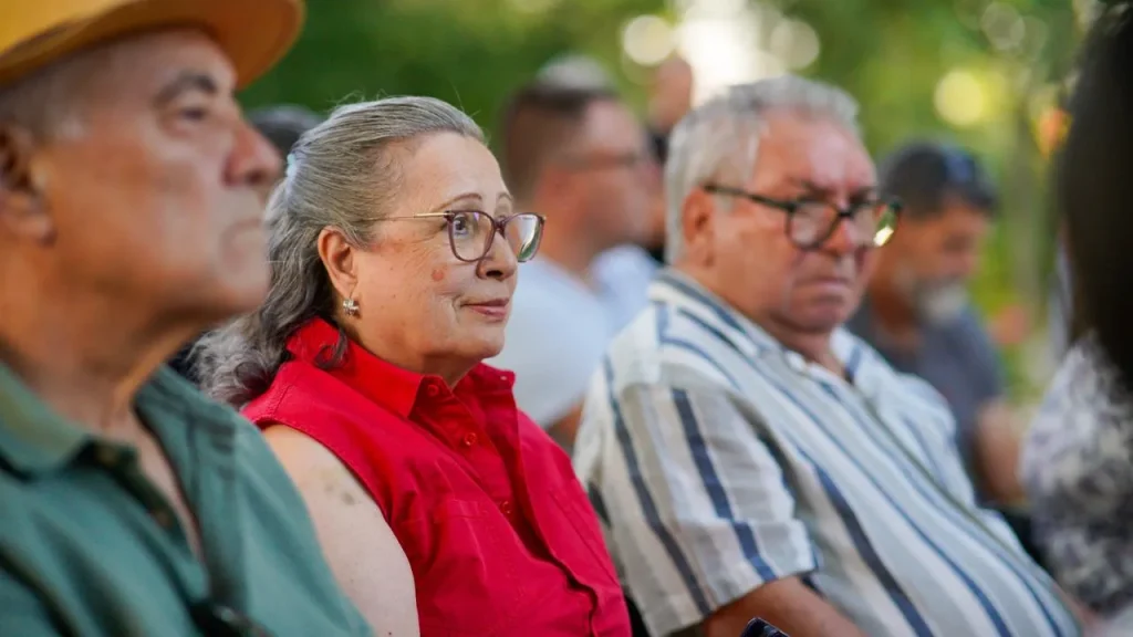 Un grupo de personas mayores está sentado al aire libre, mirando hacia delante. Aída Castillejo, con una camiseta roja sin mangas y gafas, está sentada entre dos hombres: uno lleva un sombrero amarillo y el otro, una camiseta a rayas. El fondo borroso evoca la comunidad de Rivas durante la campaña de reelección.