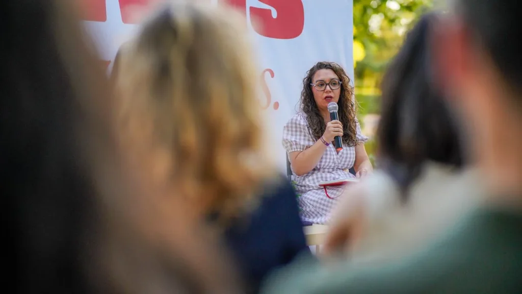 Aída Castillejo, con gafas, está sentada y habla por un micrófono en un acto al aire libre en Rivas, frente al público. La multitud, difuminada, se ve en primer plano, mientras que la vegetación ofrece un fondo lleno de vida.