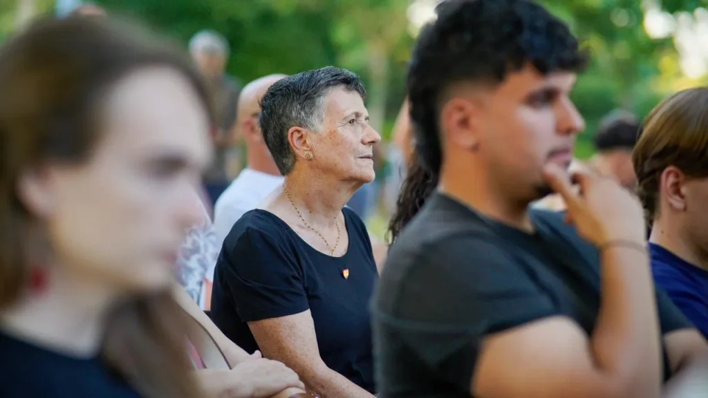 Un grupo de personas está sentado al aire libre; en el centro se encuentra Aída Castillejo, que lleva una camiseta negra y un collar, y mira hacia delante. El primer plano y el fondo están desenfocados, centrando la atención en ella, y detrás se ven árboles y Rivas.
