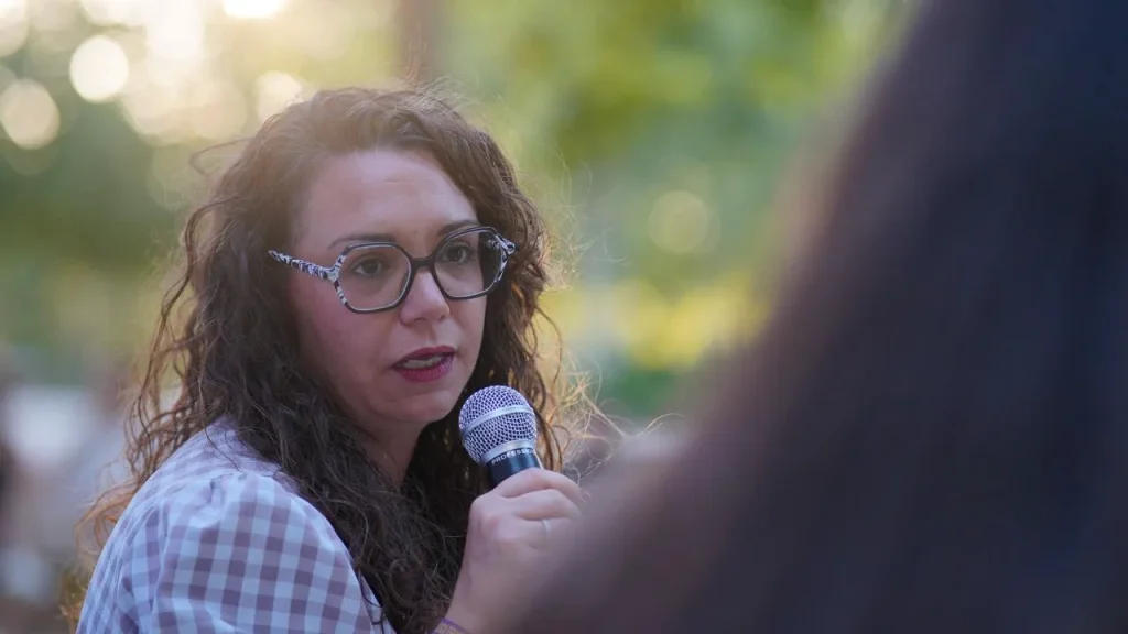 Aída Castillejo, una mujer de pelo rizado y gafas, sostiene un micrófono mientras habla al aire libre en Rivas. El fondo aparece difuminado por la vegetación y la luz del sol. En primer plano se ve parcialmente a otra persona durante el acto sobre la reelección.