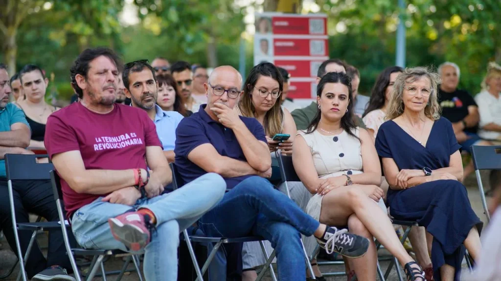 Un grupo de personas está sentado al aire libre en sillas plegables, escuchando con atención mientras habla Aída Castillejo. Al fondo se ven árboles y una pila de cajas rojas. Algunos parecen pensativos, quizá reflexionando sobre su postura respecto a la reelección.