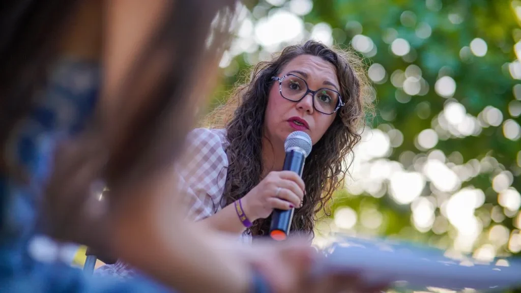 Aída Castillejo, con el pelo rizado y gafas, sostiene un micrófono y habla con atención al aire libre en Rivas. El fondo aparece difuminado por el follaje verde, y en primer plano se ve parcialmente a otra persona.