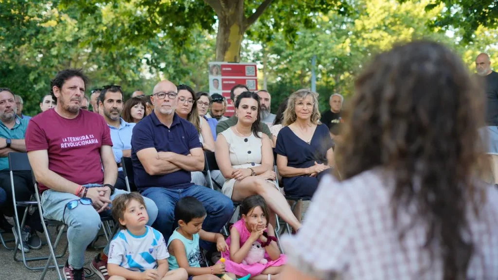 Un grupo de adultos está sentado en sillas y los niños en el suelo, escuchando a Aída Castillejo hablar al aire libre, en una zona sombreada por árboles en Rivas, mientras expone sus planes de reelección.