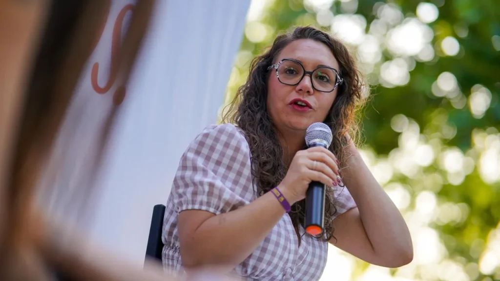 Aída Castillejo, con el pelo largo y rizado y gafas, sostiene un micrófono mientras habla al aire libre. Lleva una camisa a cuadros y está sentada frente a un fondo difuminado con vegetación en Rivas, durante su campaña de reelección.