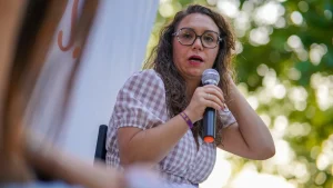 Aída Castillejo, vestida con un vestido a cuadros, está sentada al aire libre en Rivas, con el pelo largo y rizado y gafas, sosteniendo un micrófono mientras habla —con una mano tocándose el pelo— ante un fondo de árboles verdes difuminados.