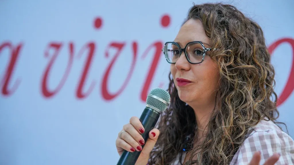 Aída Castillejo, una mujer de pelo rizado y gafas, habla por un micrófono. Lleva las uñas pintadas de rojo y está de pie frente a un fondo borroso con texto en rojo; es posible que esté hablando de temas relacionados con la reelección en Rivas.
