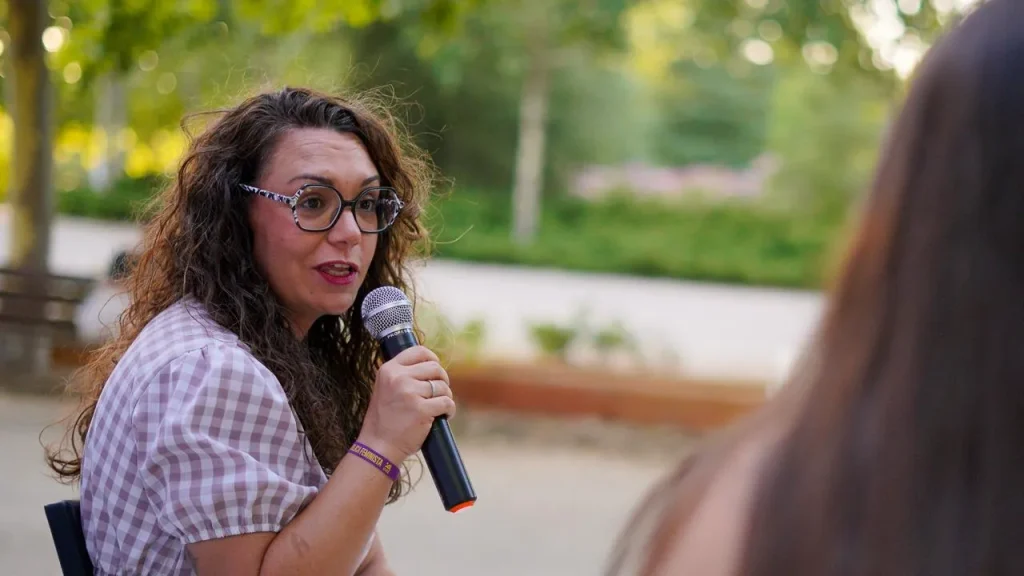 Aída Castillejo, con el pelo largo y rizado y gafas, habla por un micrófono al aire libre en Rivas. Lleva una camisa a cuadros y está sentada frente a otra persona, con árboles y vegetación al fondo.