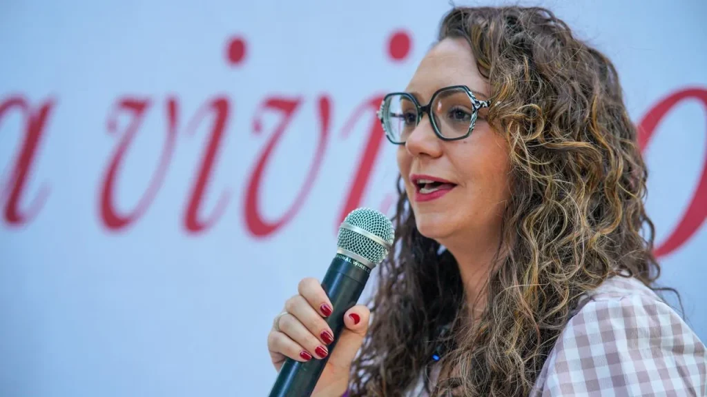 Aída Castillejo, con el pelo largo y rizado y unas gafas grandes y redondas, sostiene un micrófono y habla. Viste una camiseta clara a cuadros y se encuentra de pie frente a un fondo borroso en el que se lee, en letras rojas, la palabra «reelección».