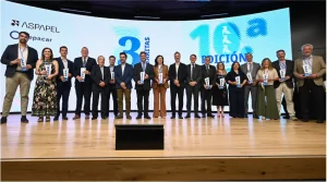 Un grupo de dieciocho personas vestidas con traje de negocios posa en un escenario con los premios en la mano, para una foto frente a una gran pantalla en la que se muestran datos del evento y los logotipos de los patrocinadores.