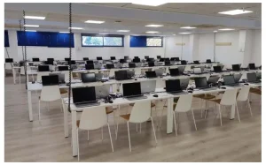 Una amplia sala con filas de mesas y sillas blancas, cada puesto equipado con un ordenador portátil, un teclado, un ratón y un monitor. Ideal para los eventos de la bolsa de empleo de Rivamadrid, la sala cuenta con persianas azules y una iluminación cenital muy luminosa.