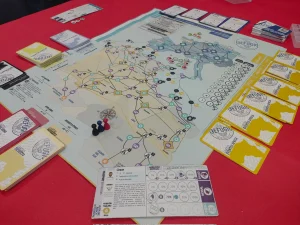 En una mesa roja de Ludiversia hay un juego de mesa con un mapa con rutas, fichas de colores, varios mazos de cartas y cartas de jugador de personaje. Varias fichas invitan a probar juegos con amigos.