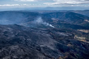 Vista aérea de un paisaje montañoso con grandes extensiones de tierra calcinada y ennegrecida y manchas de humo que se elevan desde el bosque, lo que indica los daños provocados por un reciente incendio forestal. En este borrador automático se aprecia algo de vegetación verde y un pequeño asentamiento.