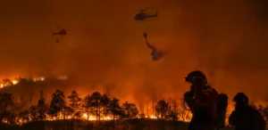 Un incendio forestal quema un bosque por la noche. Dos helicópteros con borrador automático lanzan agua sobre las llamas, mientras un bombero con equipo permanece en primer plano, con un espeso humo llenando el cielo.