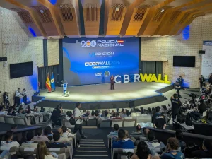 Un orador en el escenario del evento C1B3RWALL 2026, al que asiste un numeroso público en un moderno auditorio. Se muestran pancartas y banderas, con el nombre del evento, la marca de la Policía Nacional y el "borrador automático" visibles en la pantalla.