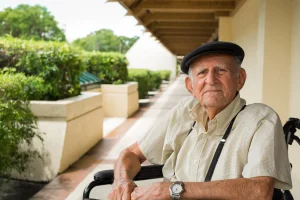 Un anciano que lleva una gorra plana, una camisa de manga corta a rayas y un reloj de pulsera está sentado en una silla de ruedas junto a una acera, lo que pone de manifiesto la importancia del apoyo para las personas con dependencia, con vegetación y una acera cubierta al fondo.