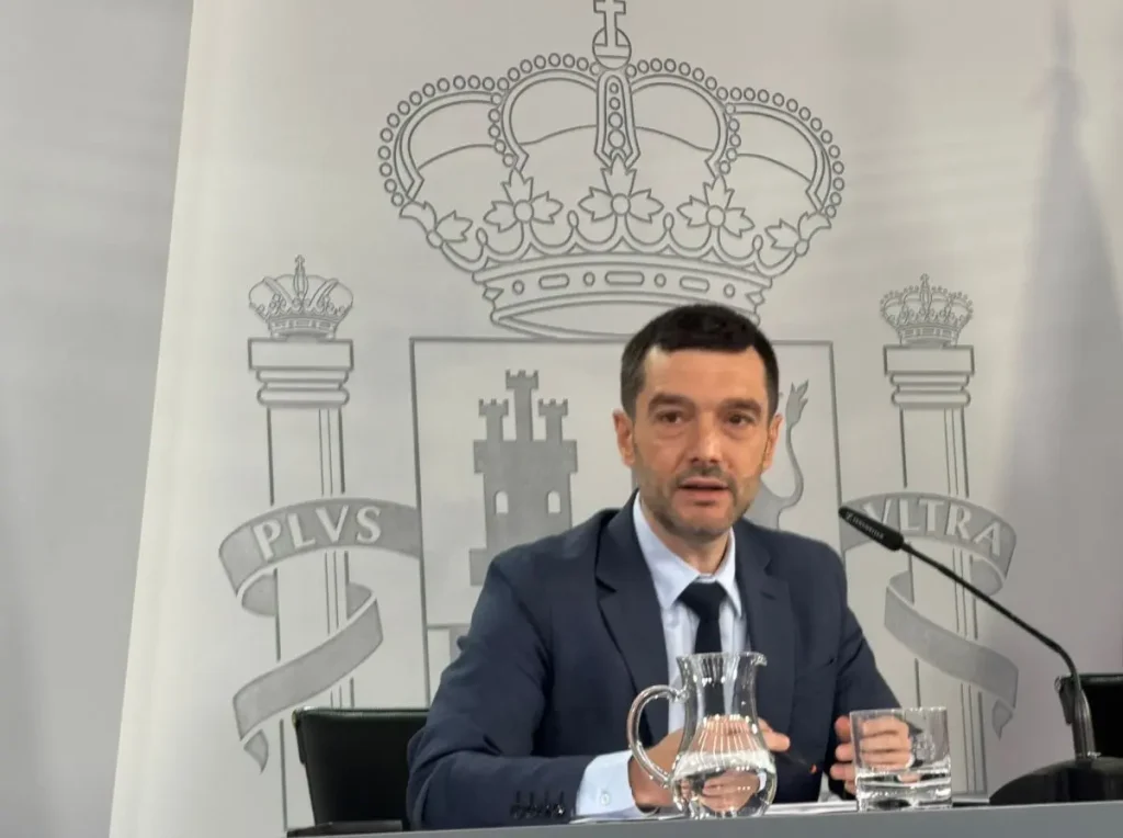 Un hombre vestido con un traje oscuro está sentado a una mesa junto a un micrófono, un vaso de agua y una jarra, listo para abordar las exigencias de mejora. Detrás de él hay un telón de fondo en el que se ve una gran corona y un escudo con el texto «PLVS VLTRA».