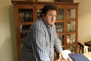 Un hombre con una camisa a rayas está de pie, apoyado en una mesa de madera frente a una vitrina llena de vajilla y cristalería; su expresión seria deja entrever la gravedad del caso Gürtel.