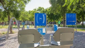 En un parque con árboles frondosos hay un conjunto de aparatos de gimnasia al aire libre instalados sobre una superficie de grava. En unos paneles informativos azules se muestran diagramas de ejercicios de «Borrador automático». Al fondo se ve a una persona con una camiseta verde.