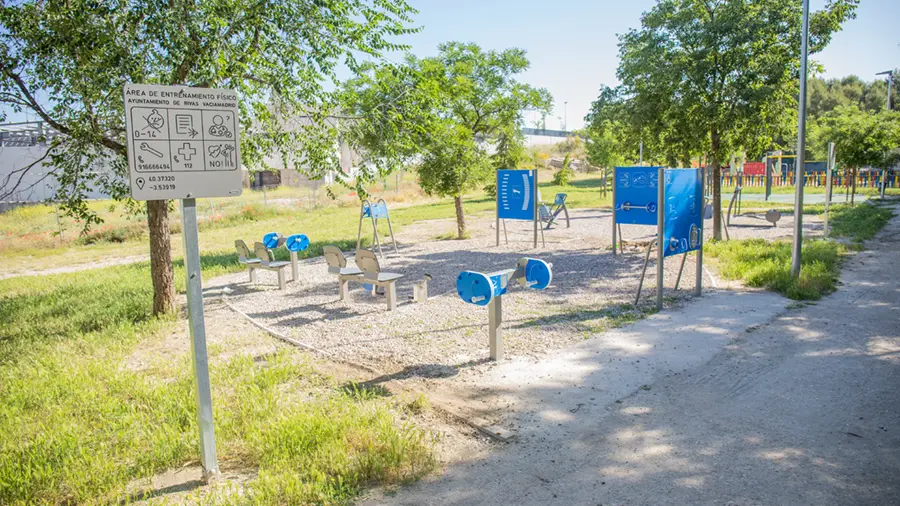 Zona de fitness al aire libre en un parque con diversas áreas biosaludables y nuevas zonas de ejercicio sobre grava, rodeadas de césped y árboles en Rivas. A la izquierda se ve un cartel con instrucciones de uso y normas de seguridad.