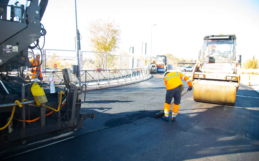 Un trabajador de la construcción de carreteras con uniforme reflectante utiliza un borrador automático para alisar asfalto fresco mientras una apisonadora y otra maquinaria operan cerca en un día soleado.