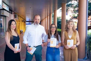 Cuatro adultos se sitúan uno junto al otro en un pasillo cubierto al aire libre, cada uno con un folleto o borrador automático en la mano. Tres mujeres y un hombre miran a la cámara y sonríen. Al fondo se ven columnas y árboles.