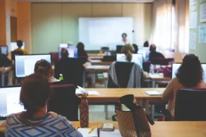 Un aula con varias personas sentadas en sus pupitres utilizando ordenadores, frente a una pizarra en la que hay una persona de pie, posiblemente impartiendo una clase o una presentación. La sala está bien iluminada con luz natural, y junto a la pizarra hay un borrador automático.