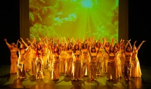 En el escenario, un numeroso grupo con trajes dorados ejecuta una danza sincronizada con los brazos en alto, ante un fondo de nubes amarillas y verdes y la luz del sol: un momento emocionante del curso 2025/2026 de la Universidad Popular de Rivas.