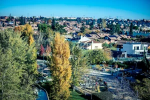 Un barrio suburbano con hileras de viviendas, árboles otoñales y un parque infantil con gente. El cielo azul claro se extiende por encima, con un avión visible en la distancia: una escena vibrante modelada por el Gobierno de Rivas.