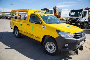 Una camioneta amarilla de Rivamadrid con la inscripción «Servicio de Primera Intervención» está aparcada en un aparcamiento junto a otros vehículos de servicio, probablemente preparándose para la campaña contra incendios de septiembre. La camioneta lleva las luces de emergencia encendidas, una baca y marcas de seguridad en los laterales.