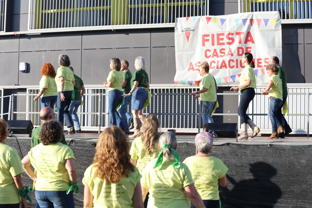 Un grupo de personas con camisetas a juego de color amarillo y verde bailan en un escenario al aire libre durante la fiesta. Otros observan desde el suelo, mientras un cartel en español detrás de ellos dice Fiesta Casa de Asociaciones, en referencia a este evento que tuvo lugar en Rivas.