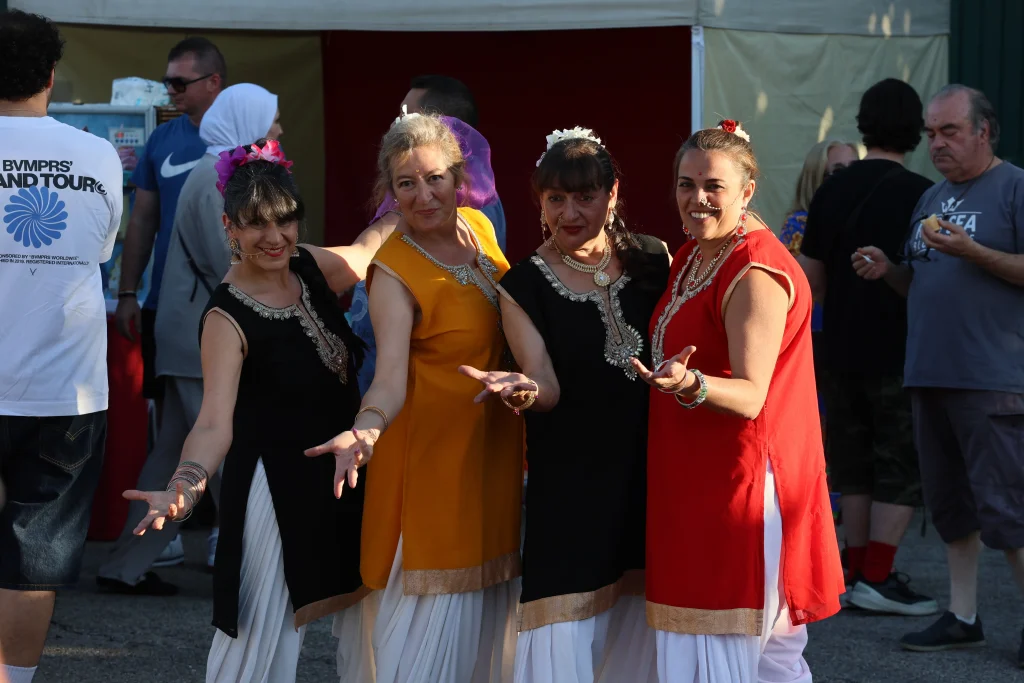 Cuatro mujeres con coloridos trajes tradicionales posan juntas al aire libre, sonriendo y gesticulando con las manos abiertas durante una animada fiesta. Al fondo, la gente se reúne cerca de las carpas en un acto para celebrar el asociacionismo ripense.
