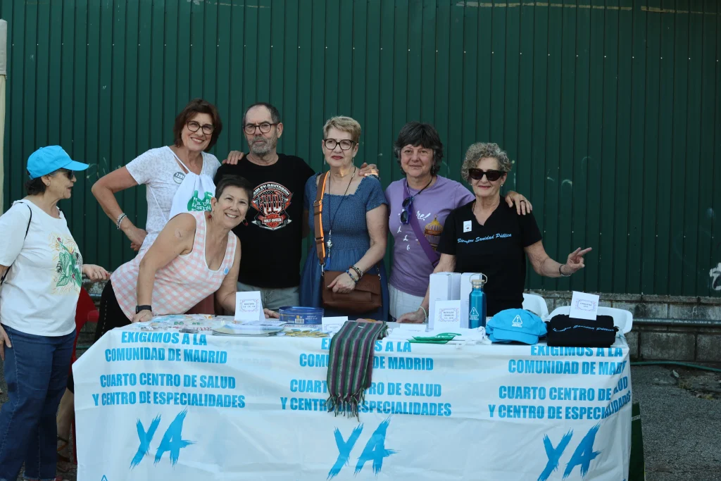 Seis adultos de pie y sentados detrás de una mesa cubierta con una pancarta blanca en español, con artículos promocionales. El grupo, que representa al asociacionismo ripense, posa al aire libre en una fiesta frente a una valla verde, algunos sonriendo a la cámara.