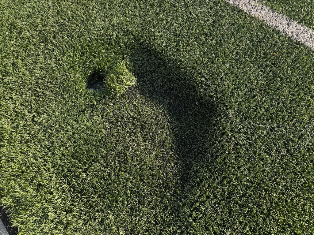 Falta un trozo de césped artificial, lo que deja al descubierto un agujero del que sobresalen algunas briznas en el campo del Cerro del Telégrafo. En el césped se aprecian parte de una línea blanca del campo y la sombra de una persona.