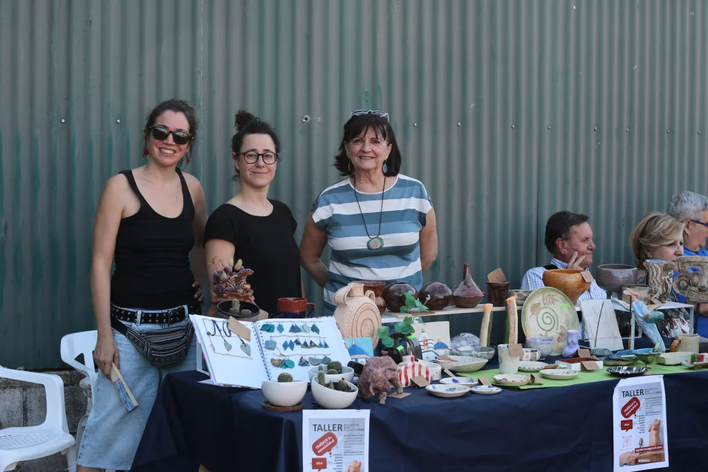 Tres mujeres de pie detrás de una mesa exponen cerámica y artesanía hechas a mano en un mercado al aire libre durante una animada fiesta, en la que el asociacionismo ripense fomenta el espíritu de comunidad. Al fondo, dos hombres charlan sentados contra un muro de chapa ondulada.