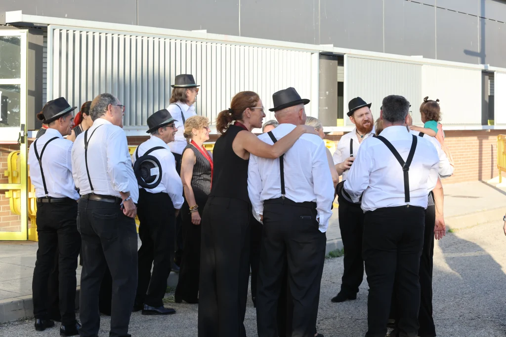 Un grupo de adultos, la mayoría vestidos con camisa blanca, tirantes negros y sombrero negro, conversan al aire libre durante una fiesta. Una mujer con coleta pasa el brazo por el hombro de un hombre. Un niño es sostenido por alguien del grupo de asociacionismo ripense.