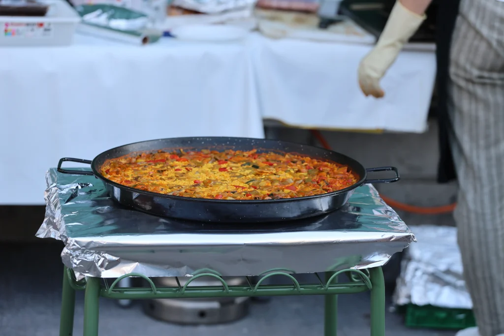 Una gran sartén negra llena de paella se encuentra sobre una mesa cubierta de papel de aluminio al aire libre, probablemente como parte de una fiesta del asociacionismo ripense. Al fondo, una persona con pantalones a rayas y un solo guante está cerca de una mesa con varios objetos.