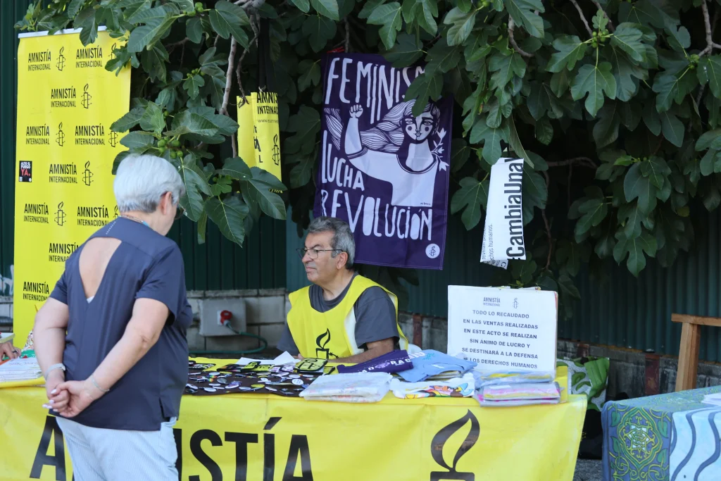 Una persona con un chaleco amarillo de Amnistía Internacional se sienta ante una mesa al aire libre cubierta de panfletos y mercancía durante una animada fiesta. Otra se encuentra cerca, con una pancarta de la Revolución Feminista y el espíritu del asociacionismo ripense de fondo.