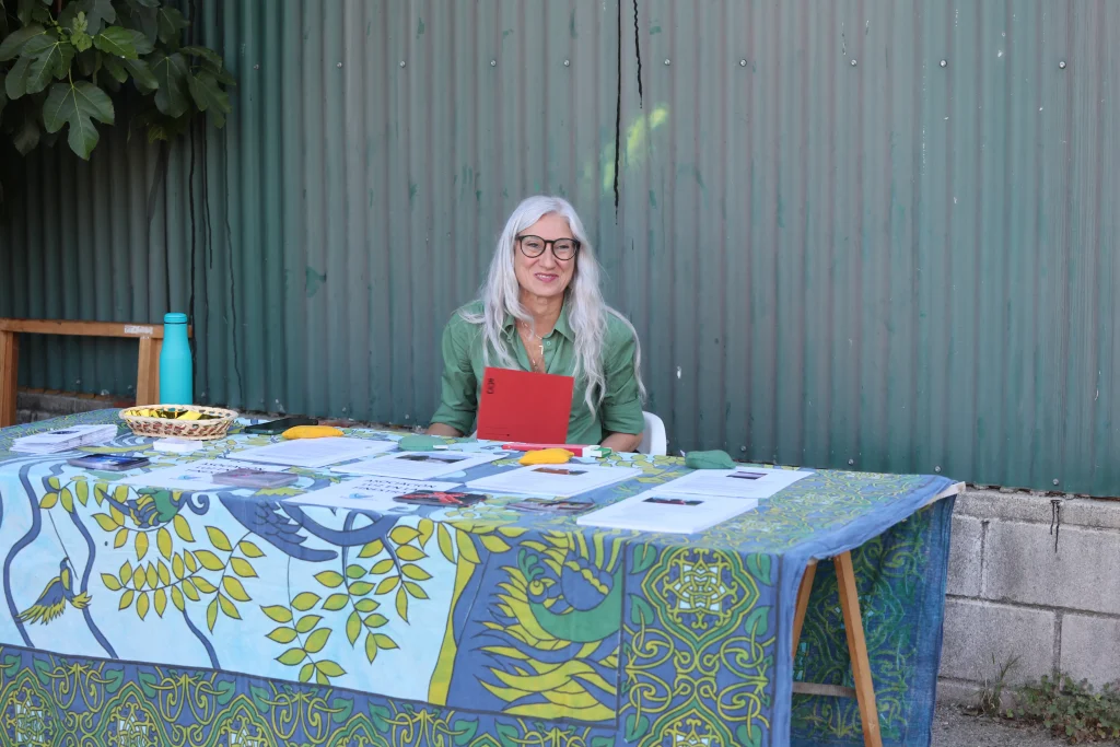 Una mujer con largas canas y gafas está sentada ante una mesa cubierta con un paño de colores, mostrando papeles y una tablilla roja, que representa el asociacionismo ripense durante la fiesta, frente a una pared de metal corrugado.