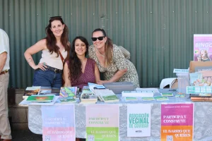 Tres mujeres de pie y sentadas detrás de una mesa con libros, folletos y carteles que promueven el asociacionismo ripense durante la fiesta. La mesa muestra folletos de colores, con una pared de chapa ondulada al fondo.