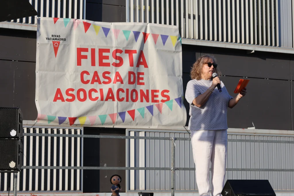 Una mujer con gafas de sol habla por un micrófono en un escenario exterior de la fiesta, que celebra el asociacionismo ripense. Detrás de ella, una pancarta con banderas de colores reza Fiesta Casa de Asociaciones, y una pequeña muñeca se sienta en el escenario.