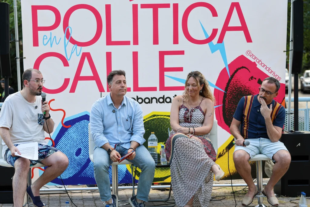 Cuatro personas están sentadas en taburetes frente a un cartel que dice «POLÍTICA en la CALLE» en Rivas. Una de ellas sostiene un micrófono y unos papeles, mientras las demás escuchan y debaten sobre el modelo de ciudad. Sobre la mesa que tienen delante hay botellas de agua y un ventilador.
